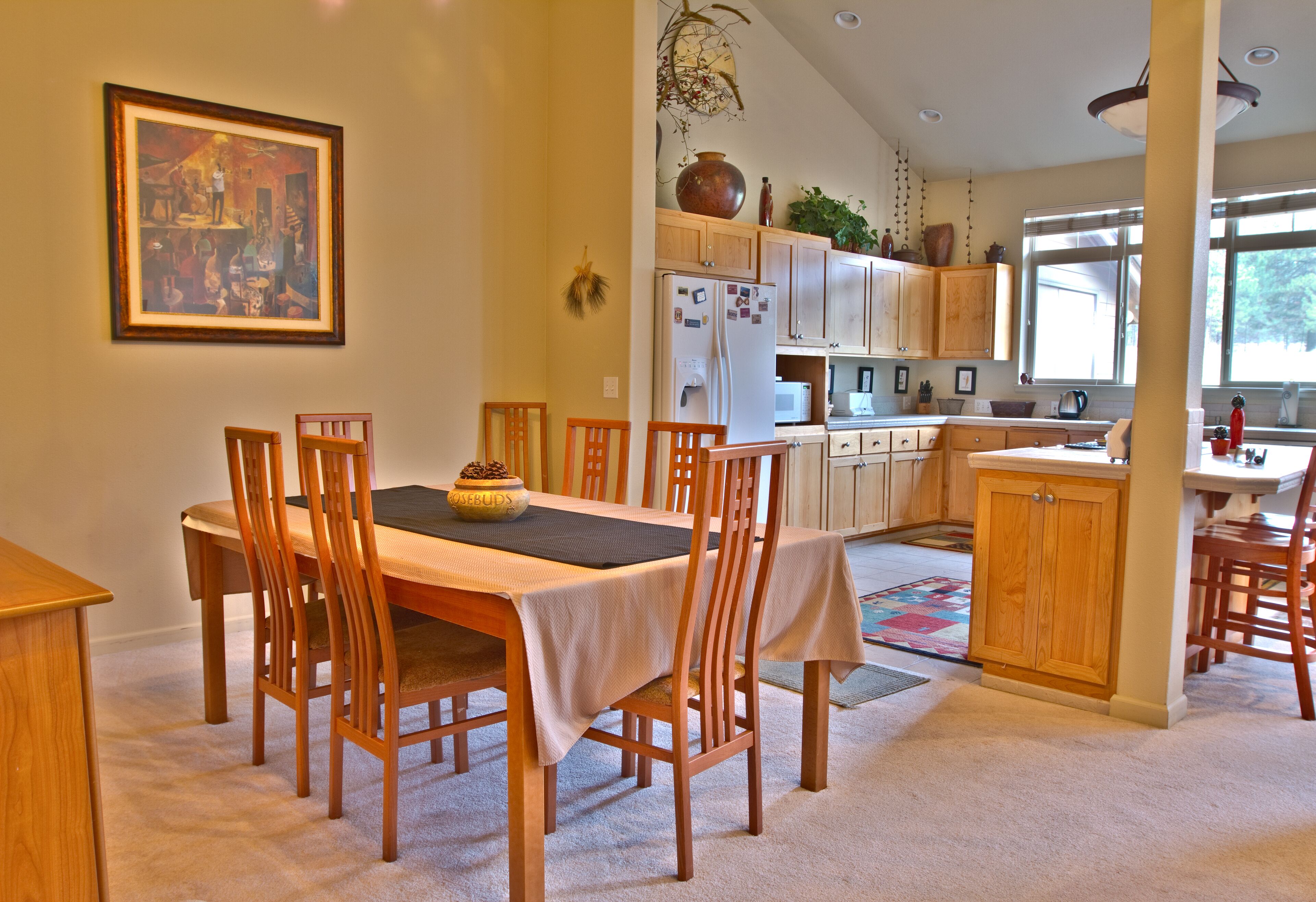 A dining area with a table set for four, wooden chairs, and a kitchen in the background with cabinets and appliances.