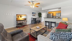 A modern living room with a flat-screen TV, a ceiling fan, a dining area, and a kitchen.