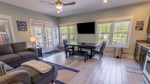 A modern living room with a sofa, coffee table, and a dining area with a table and chairs. There is a TV mounted on the wall, and a kitchen area is visible in the background. - Pet Friendly Resort Charmer w/ Golf Cart (Michigan City)