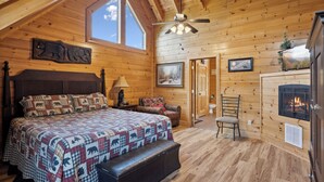 Une chambre confortable dans un chalet en rondins, avec un grand lit, une cheminée, un ventilateur de plafond et une fenêtre donnant sur le ciel.