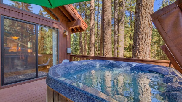A hot tub on a wooden deck with a glass door leading to a room, surrounded by tall trees.