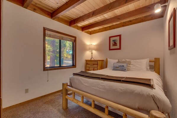 Un dormitorio con estructura de cama de madera, ventana con vista a árboles, mesita de noche con lámpara y un cuadro en la pared.