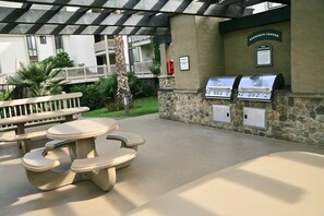 An outdoor barbecue area with stone walls, a grill, and picnic tables. - Ocean View-Close to Beachfront Cottages. E105 North Coast Village (Oceanside)