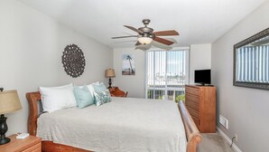 A bedroom with a bed, a ceiling fan, a dresser, a TV, and a window with a view.