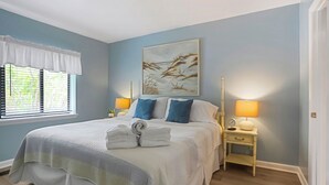A bedroom with a bed, two bedside tables with lamps, a window with blinds, and a painting on the wall. - Surf Court 70 - Forest Beach Townhouse (Hilton Head Island)