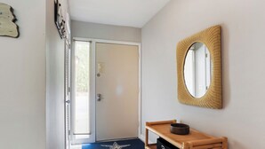 A hallway with a door, a star-patterned mat, a wicker mirror, and a wooden bench with a basket. - Surf Court 70 - Forest Beach Townhouse (Hilton Head Island)