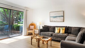 A living room with a large sliding glass door leading to a garden, a dark brown sofa with two cushions, a wicker chair, and a small table. - Surf Court 70 - Forest Beach Townhouse (Hilton Head Island)