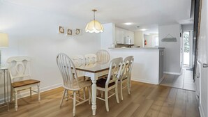A dining area with a wooden table and white chairs, a hanging light fixture, and a kitchen in the background. - Surf Court 70 - Forest Beach Townhouse (Hilton Head Island)