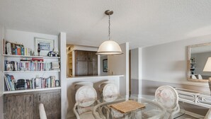 Et spiseområde med et glasbord omgivet af fletstole, en reol med bøger, et træskab og en hængende pendel.