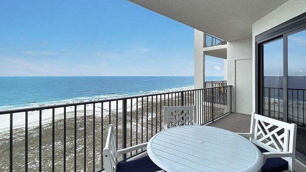 Ein Balkon mit weißem Tisch und Stühlen, der Blick auf einen Strand mit Wellen und einem klaren Himmel bietet.