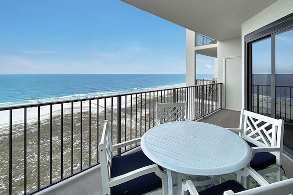 Un balcone con un tavolo e sedie bianche, vista su una spiaggia con onde e cielo sereno.