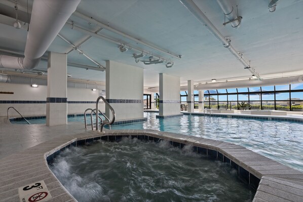 A large indoor swimming pool with a hot tub area, clear ceiling pipes, and a view of the outdoors through large windows.