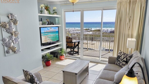 A living room with a large TV, a sofa, a coffee table, and a balcony with a view of the beach. - Summer Place 207 - 3 BR Beach Condo - 2 Sets of Beach Service! (Fort Walton Beach)