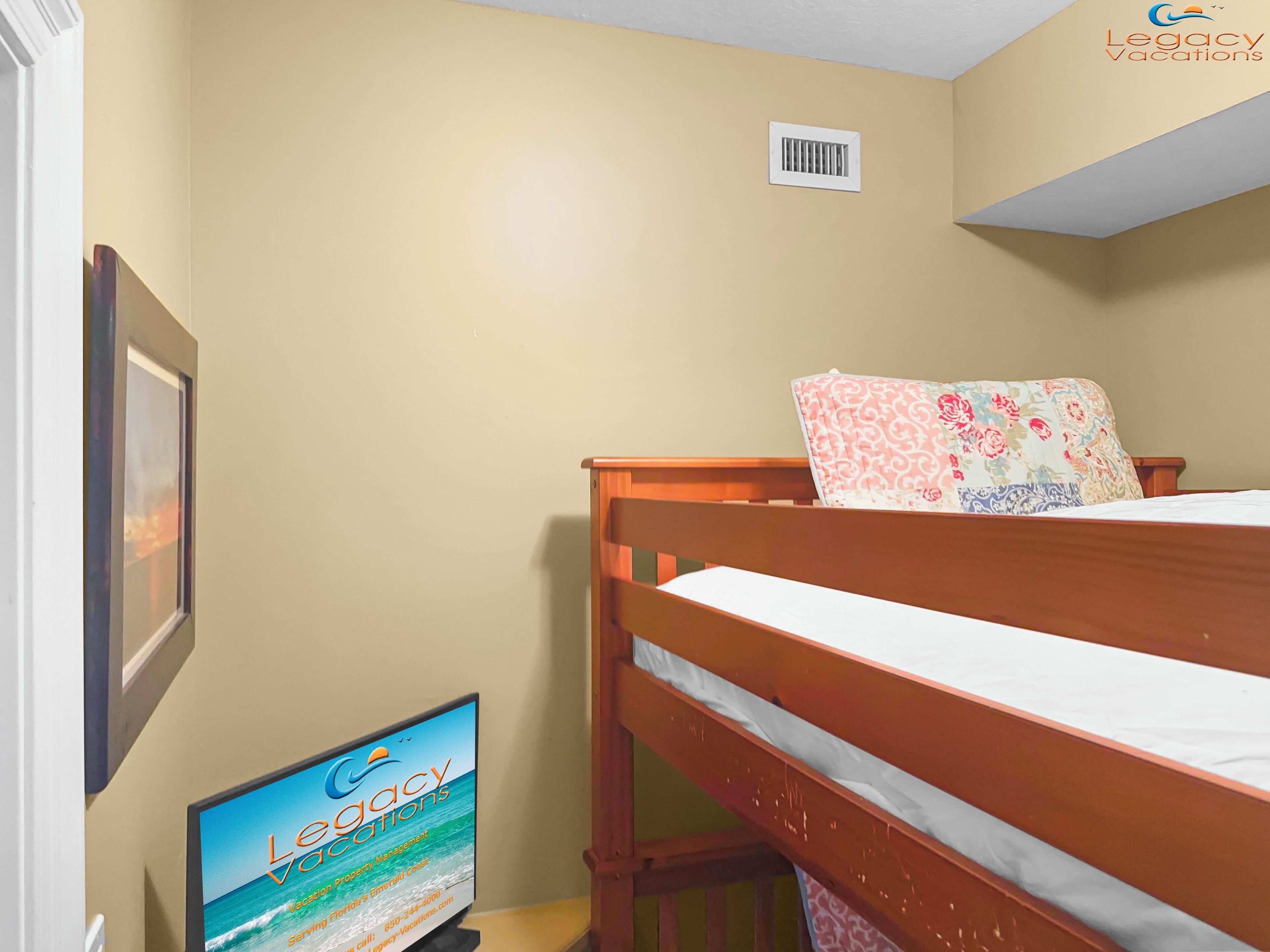 A bedroom with a wooden bunk bed, a framed picture on the wall, and a flat-screen TV displaying 'Legacy Vacations'.
