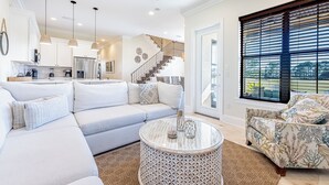 A modern living room with a sofa, a patterned armchair, a round coffee table, and a view of the kitchen and staircase.