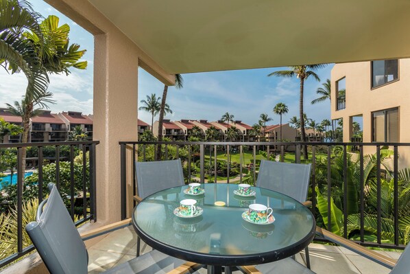A glass table with four teacups and saucers, surrounded by chairs, on a balcony with a view of palm trees and buildings.