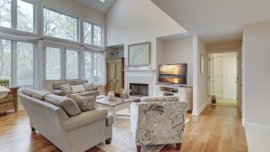 A spacious living room with a fireplace, a television, and a large window overlooking trees. - 69 Shell Ring - Nestled in A Quiet Sea Pines Community offering Pool & Tennis (Hilton Head Island)