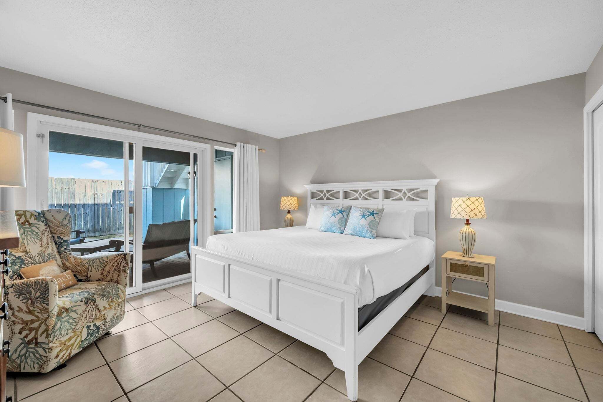 A bedroom with a white bed, a chair, a nightstand, and a view through a sliding door.