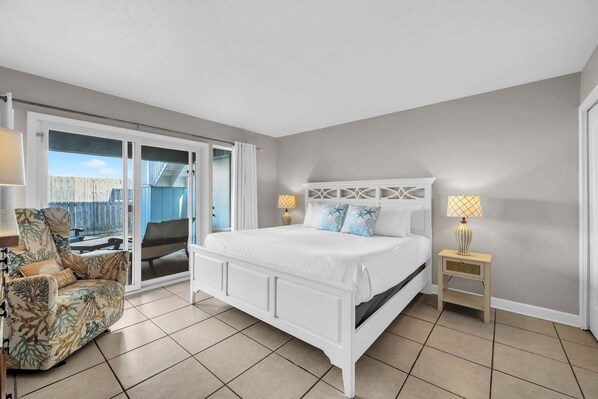 A bedroom with a white bed, a chair, a nightstand, and a view through a sliding door.