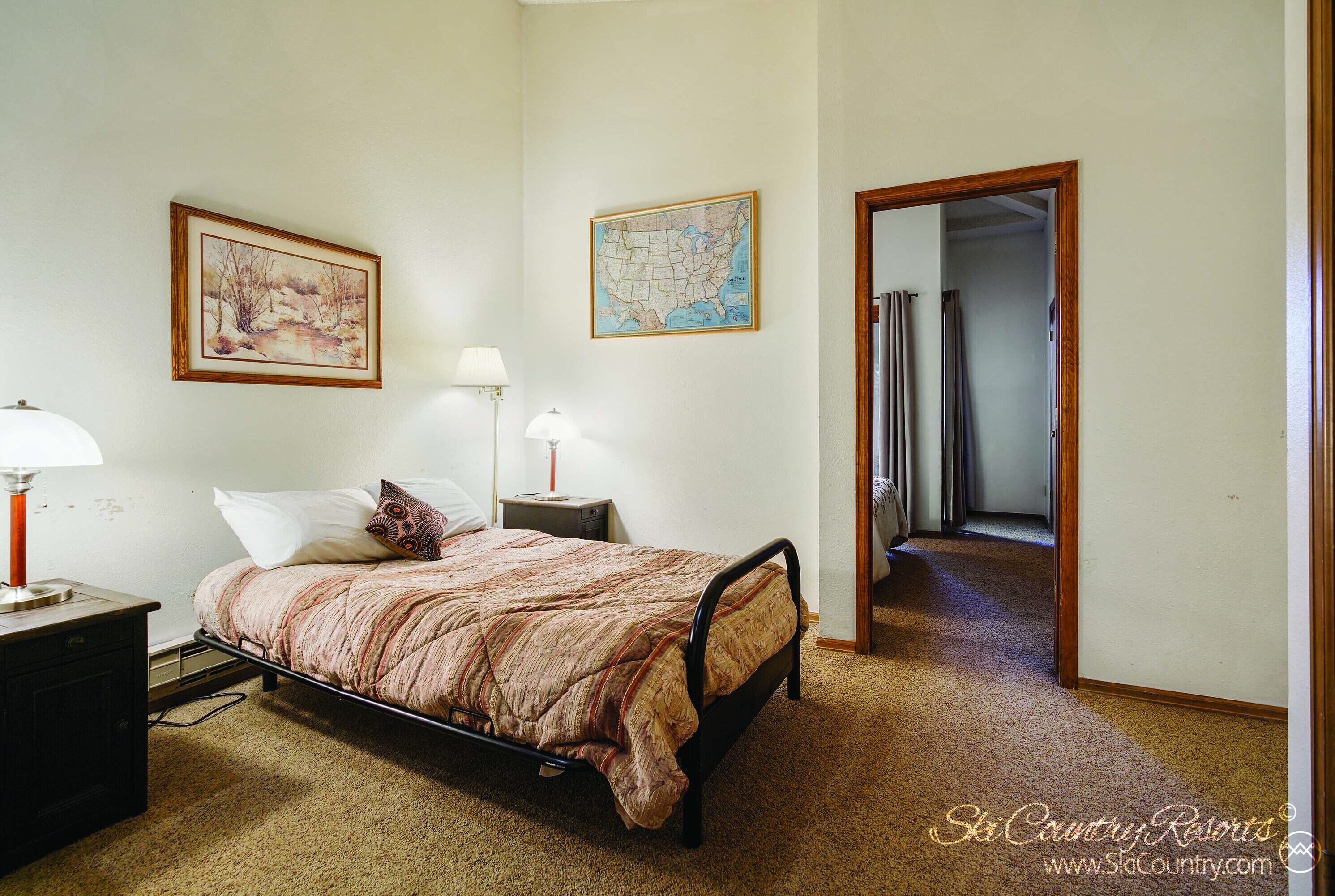 A bedroom with a bed, two bedside tables, a lamp, a map on the wall, and a door leading to another room.