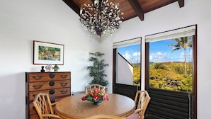 Een eethoek met een houten tafel en stoelen, een kroonluchter, een plant en uitzicht op een landschap door een raam.