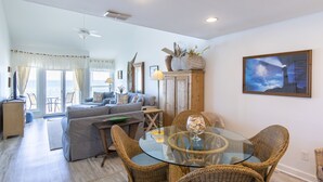 A spacious living room with a glass-top dining table surrounded by wicker chairs, a grey sofa, and a wooden cabinet with decorative items. - Gulf Front Condo, 3rd level, Amazing Views - Near shops/restaurants (Santa Rosa Beach)
