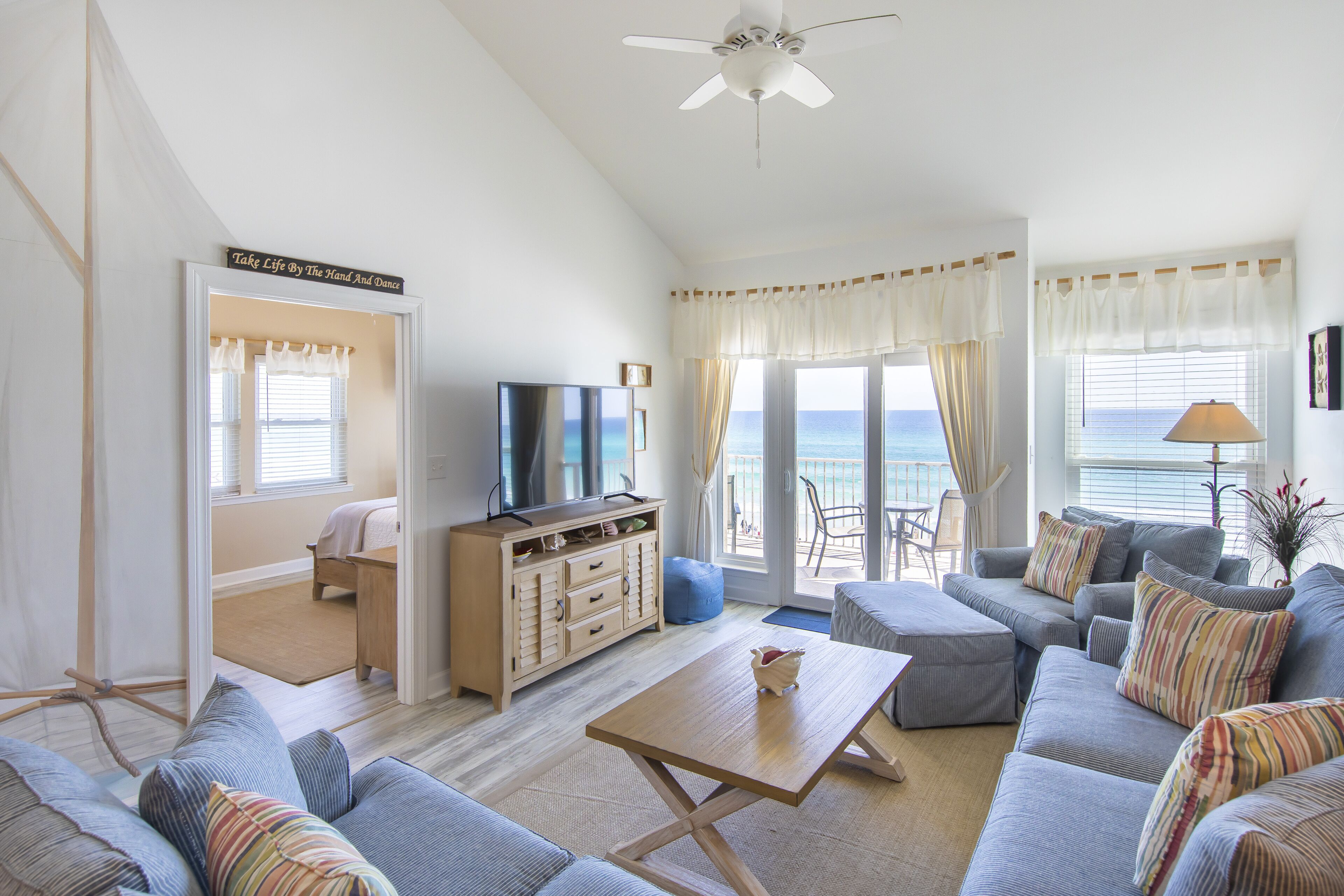 Ein geräumiges Wohnzimmer mit einer großen Couch-Garnitur, einem Couchtisch und einem Fernseher. Durch die Fenster hat man einen Blick auf das Meer.