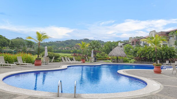 A swimming pool with lounge chairs and potted plants, surrounded by a landscaped area and buildings in the background. - Luxurious Ground Floor Condo: Proximity to Pool, Full Access to Amenities. (Herradura)