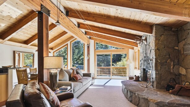 Un salón acogedor con chimenea de piedra, vigas de madera y un ventanal grande con vistas a un bosque.