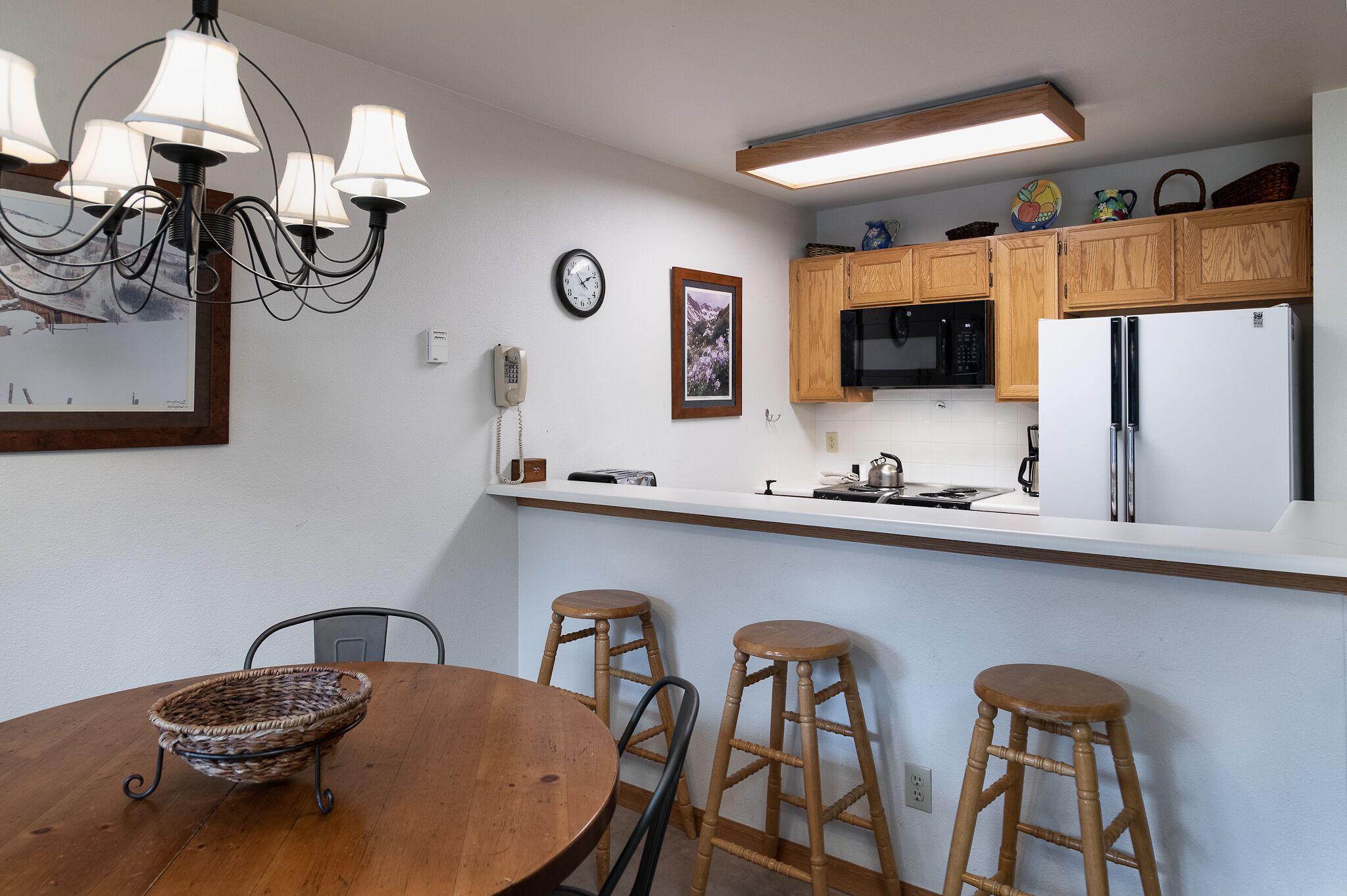 Een keuken met houten kasten, een koelkast, een magnetron en een kroonluchter. Er is een eettafel met krukken en een mandje.