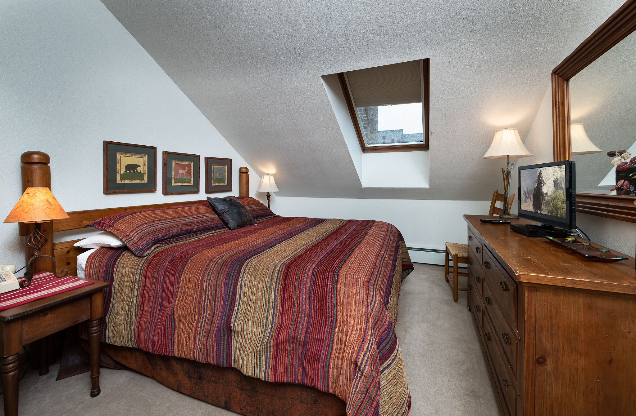 A bedroom with a large bed, a desk with a lamp, a dresser, and a television. There are framed pictures on the wall and a window with a view of buildings.