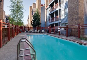 Una piscina con una escalera metálica, rodeada de barandillas rojas, y contigua a un edificio con balcones y fachada de ladrillo.