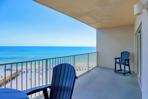 A balcony with a view of the beach and ocean. There are two Adirondack chairs and a table.