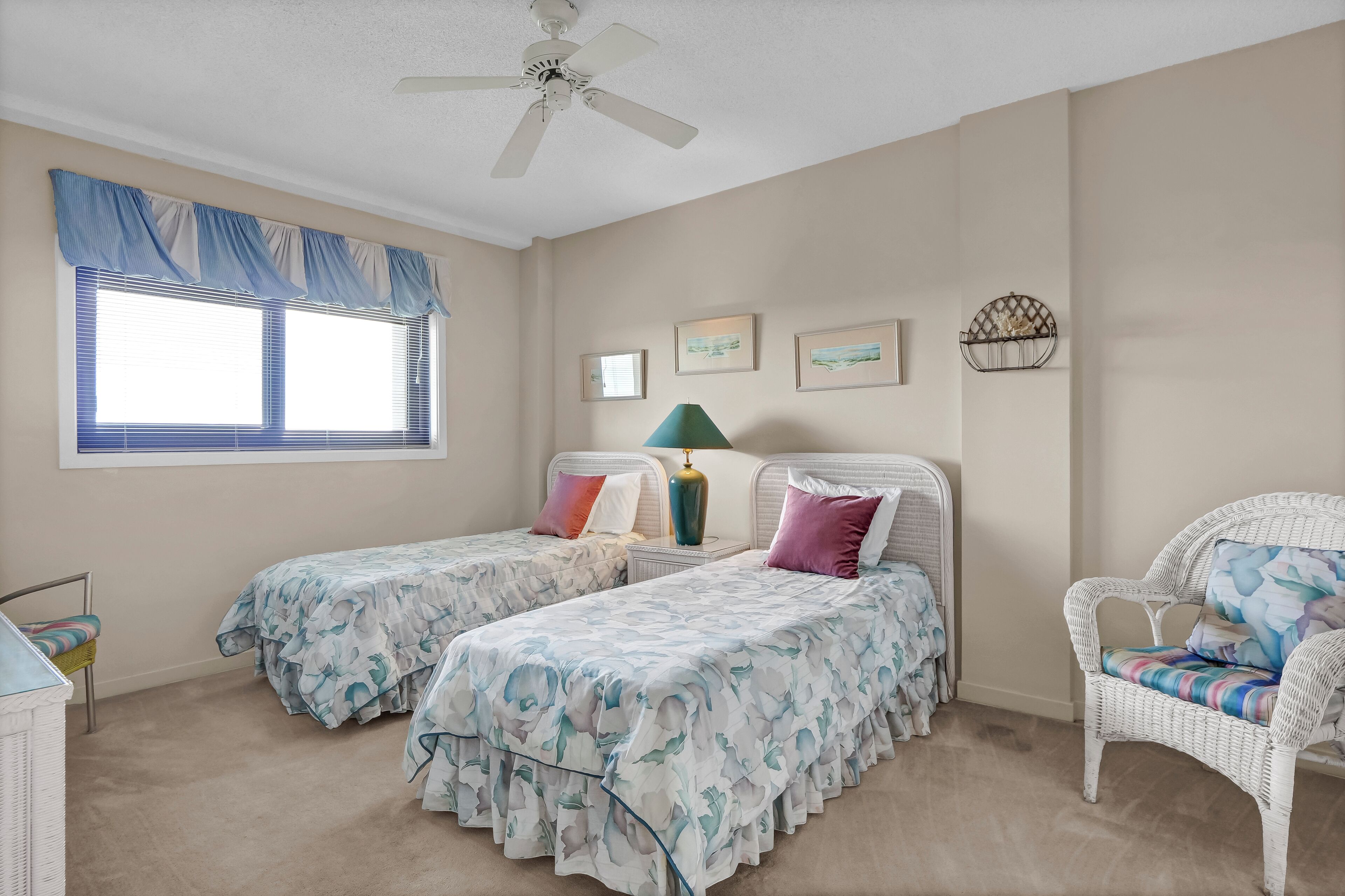 A bedroom with two beds, a window with blinds, a ceiling fan, and a wicker chair.