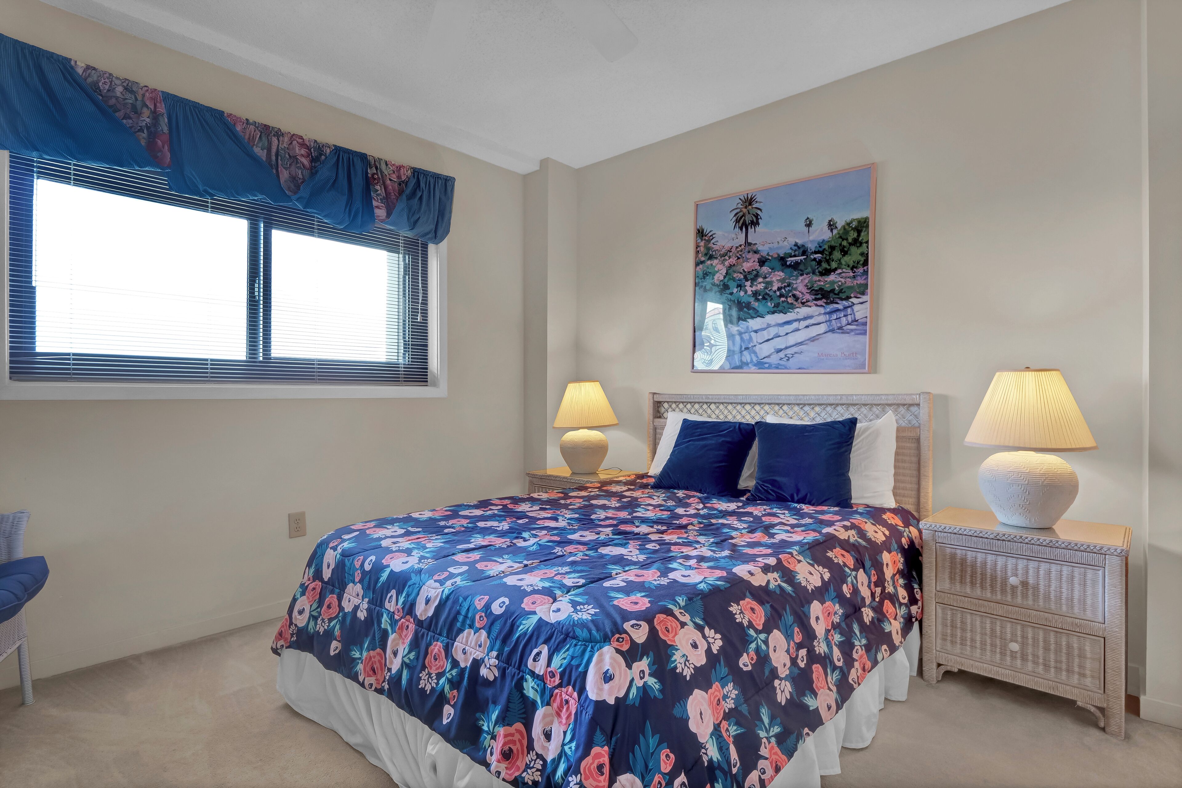 A bedroom with a large bed, two bedside lamps, a nightstand, a window with blinds, and a painting on the wall.