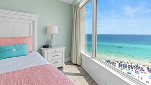 Un dormitorio con un ventanal que da a una playa, una cama con ropa de cama rosa, una mesita de noche con una lámpara y un aparador.