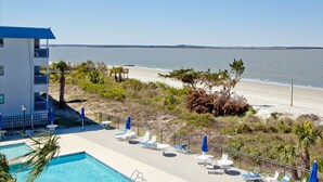 Ein Poolbereich mit Liegestühlen und Sonnenschirmen, Blick auf einen Strand und ein Gebäude mit blauem Dach.