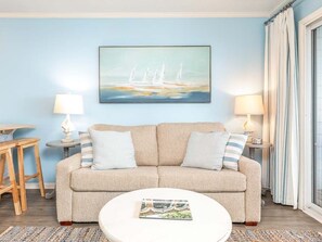 A living room with a beige sofa, two side tables with lamps, a round coffee table with a magazine, and a painting of sailboats on the wall.