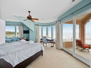 A bedroom with a large bed, a ceiling fan, a dining area with a table and chairs, and a balcony with a view of the beach.