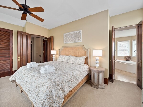 A bedroom with a large bed, two bedside lamps, a ceiling fan, a wooden headboard, a wall-mounted artwork, and a bathroom with a bathtub and window.