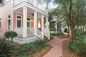 Una casa rosa con un portico bianco, un vialetto in mattoni e vegetazione circostante.