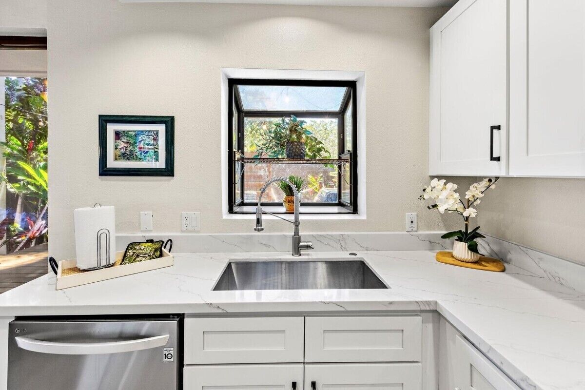 A modern kitchen with a built-in sink, white cabinetry, a window with a view of greenery, and a framed picture on the wall.