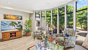A spacious living room with large windows, a comfortable seating area, and a flat-screen TV on a wooden cabinet. - The Residences at Napili Bay 8  Townhome In Lush Tropical Setting (Lahaina)