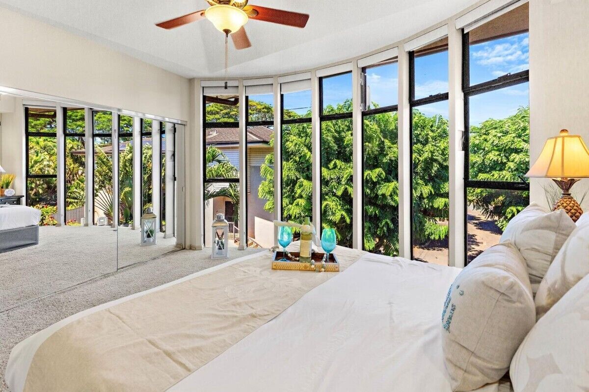 A spacious bedroom with a large bed, a ceiling fan, and a view of lush greenery through large windows.