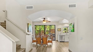 A spacious dining area with a high ceiling fan, a staircase on the left, and a kitchen area visible through a glass door. - The Residences at Napili Bay 8  Townhome In Lush Tropical Setting (Lahaina)