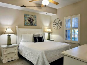 A bedroom with a bed, two bedside tables with lamps, a ceiling fan, a wall painting, a compass decoration, and a window with blinds.