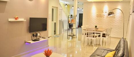 A modern living room with a flat-screen TV, a sofa, a dining area with a table and chairs, and a staircase leading to another level.