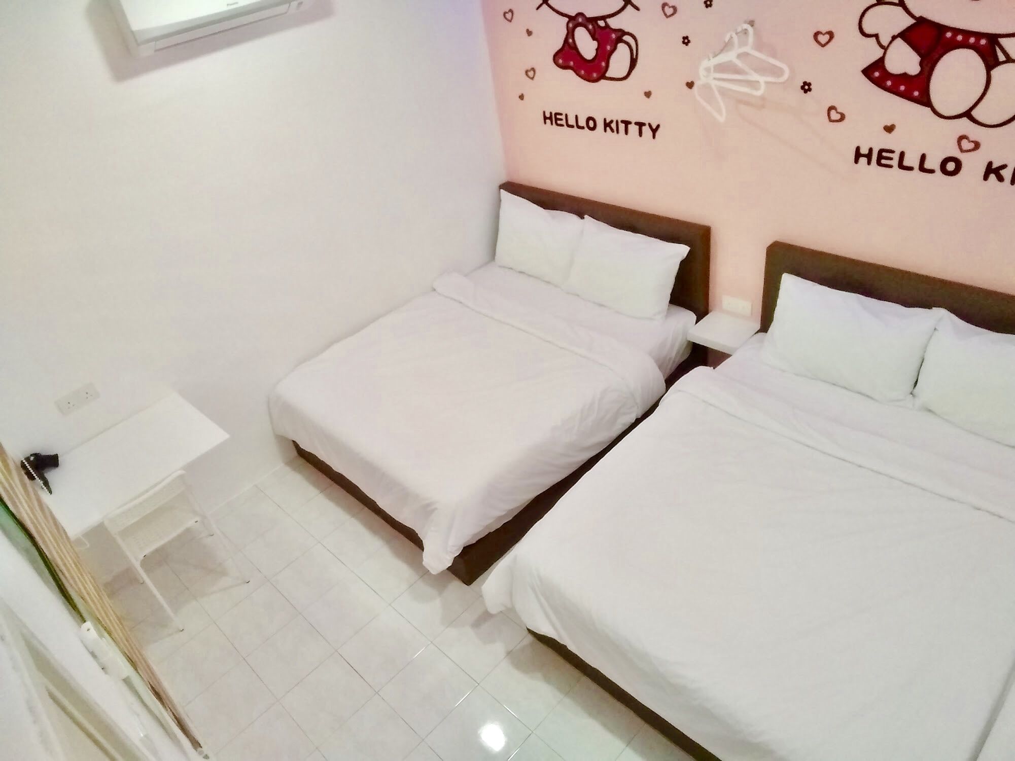 A hotel room with two single beds, a small desk, and a wall decoration featuring a cat and the text "HELLO KITTY".