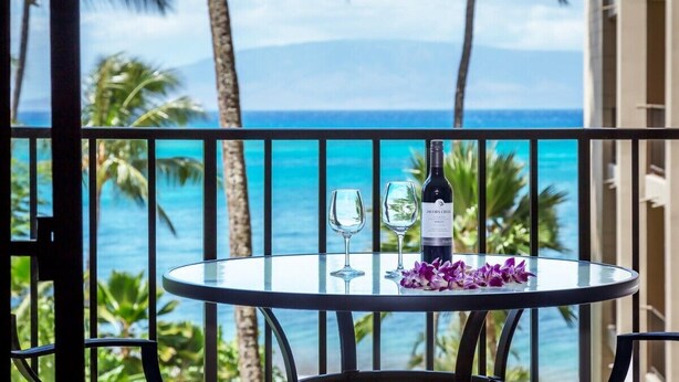 Mesa redonda con una botella de Jacob's Creek, dos copas de vino y flores moradas en un balcón con vistas a una playa tropical, palmeras y el océano.
