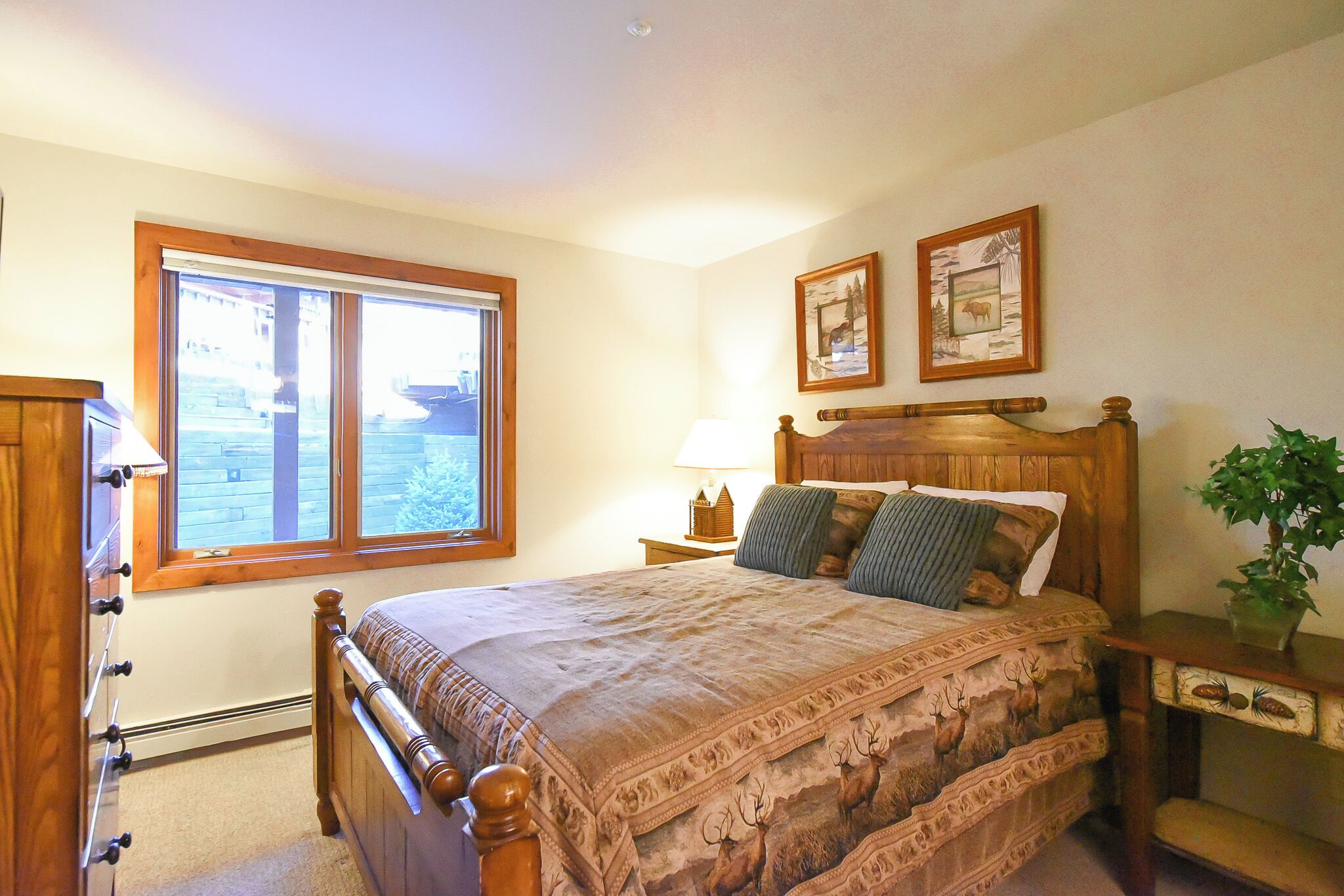 Un dormitorio con cama de madera, una ventana, una mesita de noche y dos cuadros enmarcados en la pared.